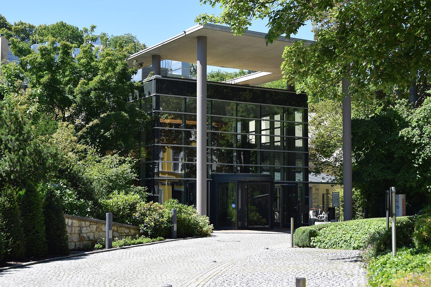 Eingang der Carolus Thermen in Aachen: gläserner Kubus unter einem weit auskragenden Betondach, davor Kopfsteinpflaster und dichtes Grün