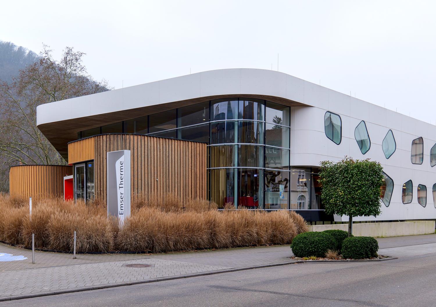 Die Emser Therme in Bad Ems von außen: geschwungener weißer Baukörper mit Holzfassade und runden Fenstern, davor Ziergräser