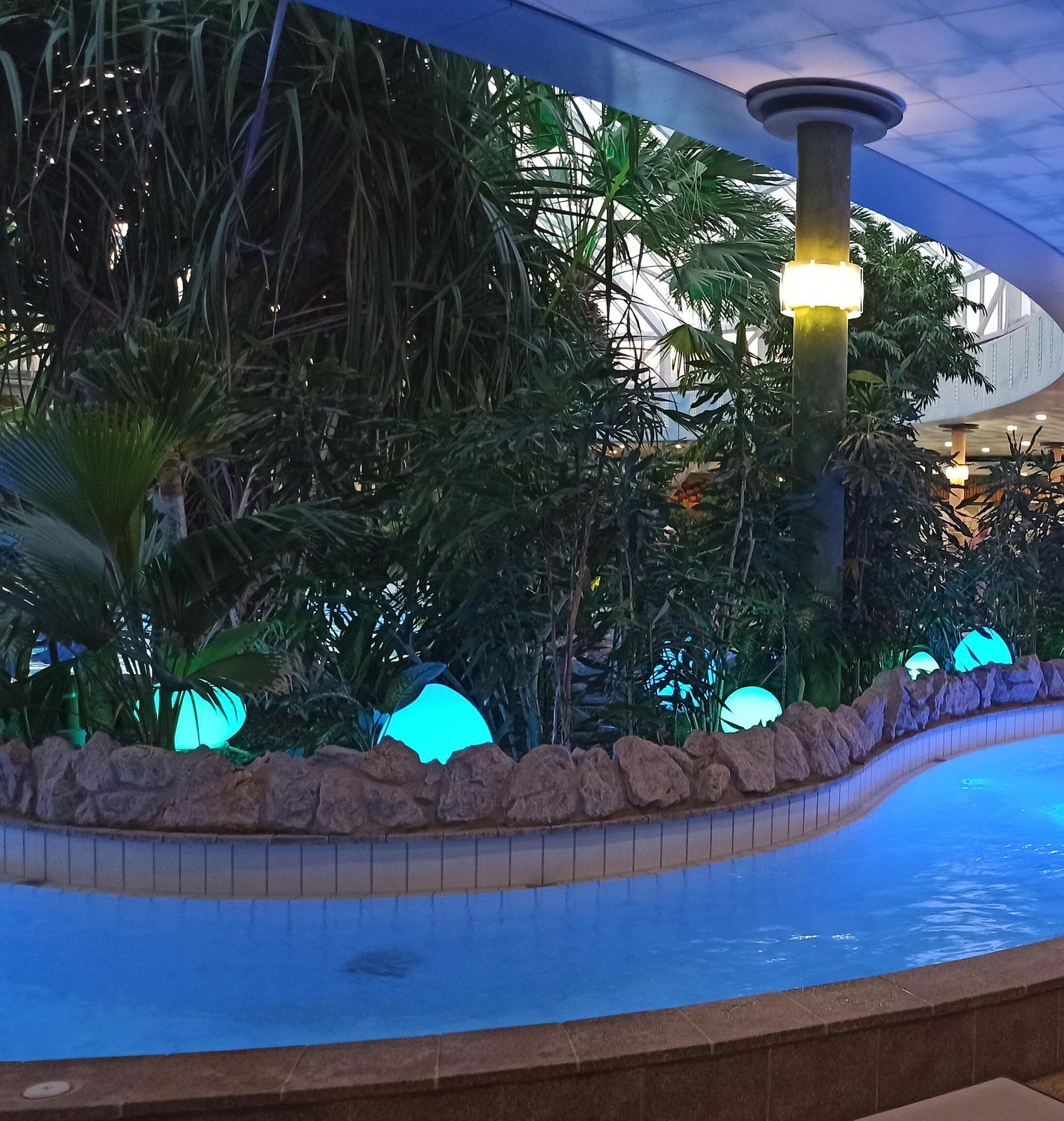 Therme Erding innen: blau beleuchtetes Becken, am Rand Felsen, Palmen und leuchtende Kugeln unter der Hallendecke